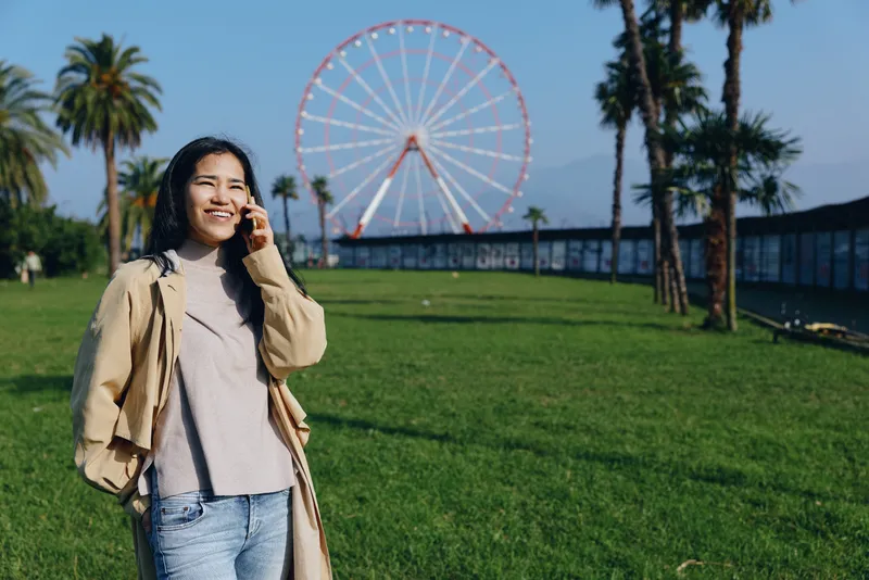 Woman standing on a sunny park lawn with a large ferris wheel in the background, smiling and relaxed in casual jacket and jeans, capturing a candid outdoor leisure moment