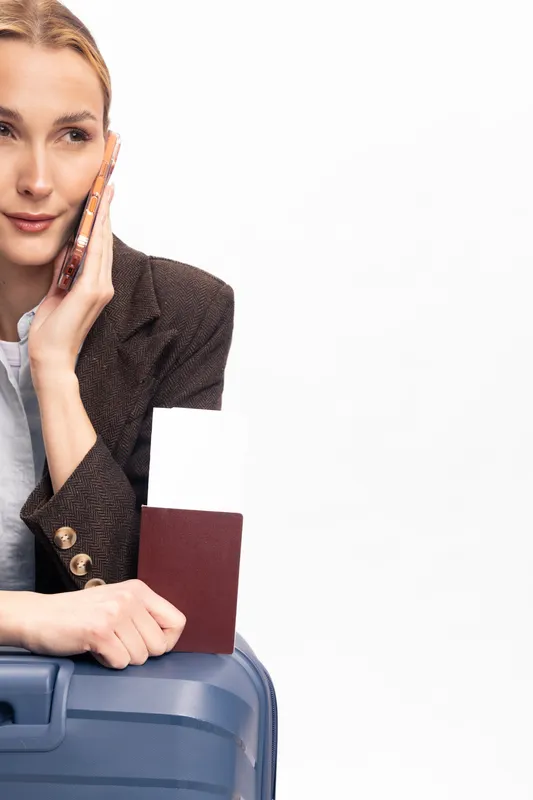 Young woman with suitcase and passport talks on smartphone. Business traveler in casual jacket looks away with thoughtful expression on white background. Travel concept with luggage and documents.