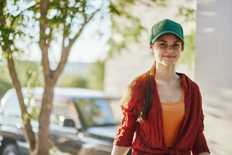 Young smiling woman in casual summer outfit, wearing a cap, walking outdoors with green background, conveying a cheerful and relaxed vibe