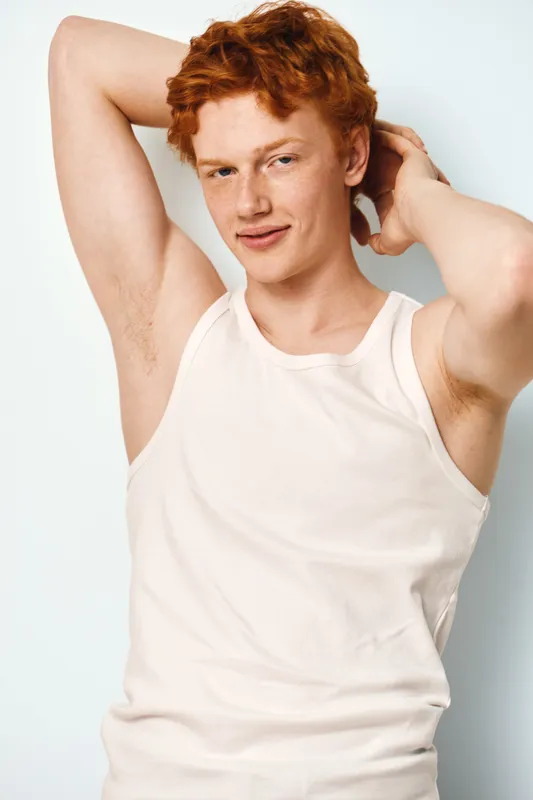 Man with red hair wearing white tank top poses with hands behind head against white background, smiling slightly, showing confident casual style and relaxed mood.