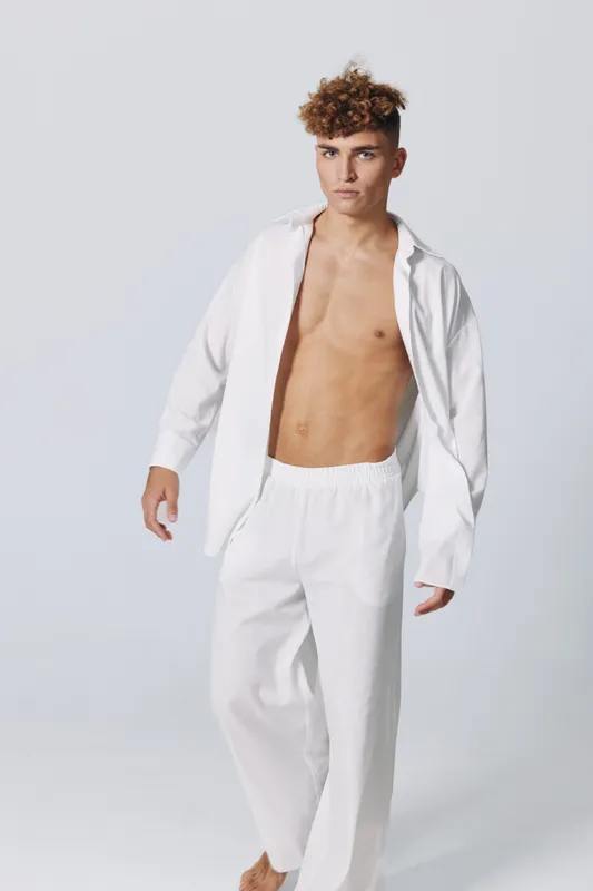 Young man with curly hair wearing white loose pants and open white shirt standing barefoot on light background. Fashion model posing with serious expression and casual style.