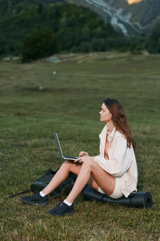 A focused young woman sits on a grassy field with a laptop, enjoying outdoor work in a calm rural setting, creating a productive yet relaxed mood for modern tasks.