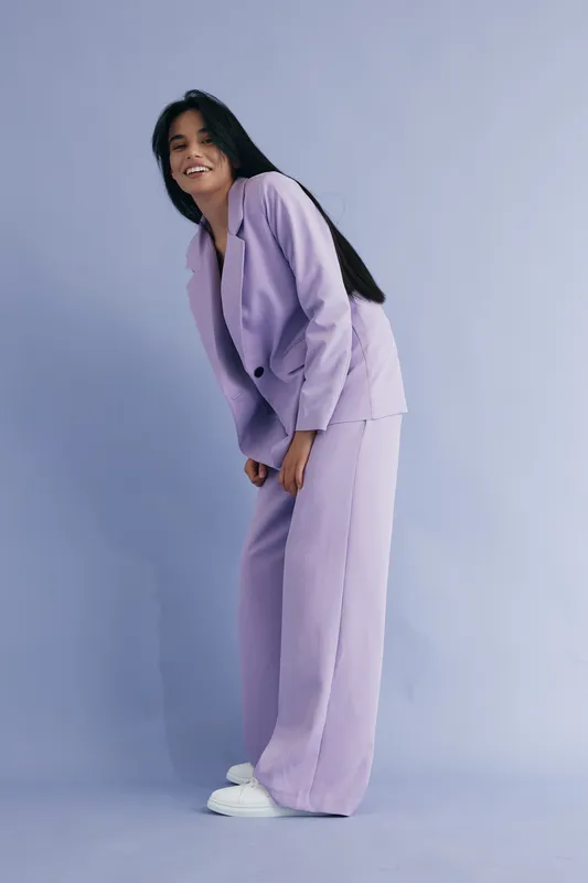 smiling woman in lilac pantsuit with long black hair standing against soft purple background, wearing white sneakers, joyful expression and casual pose