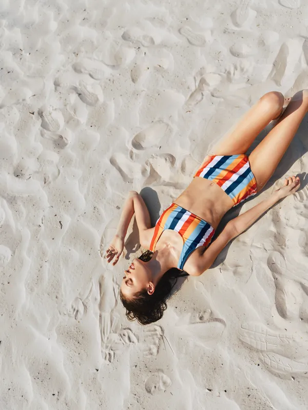 A young woman lies on sunlit sandy beach wearing a bright striped bikini, a relaxed pose, and a warm glow that emphasizes sand texture and a carefree summer mood.