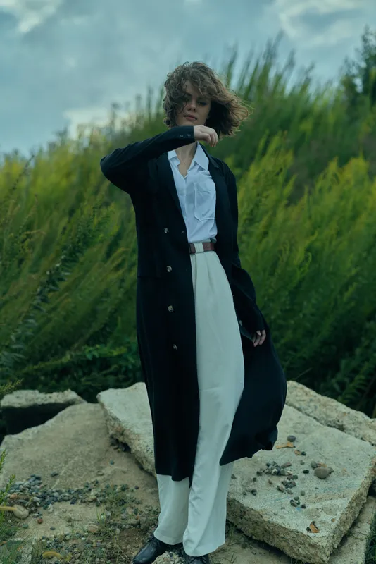 Coat, fashion, outdoor model wearing a long black coat over a white shirt and white pants on rocky terrain, windy day, showcasing confident style and contemporary outerwear
