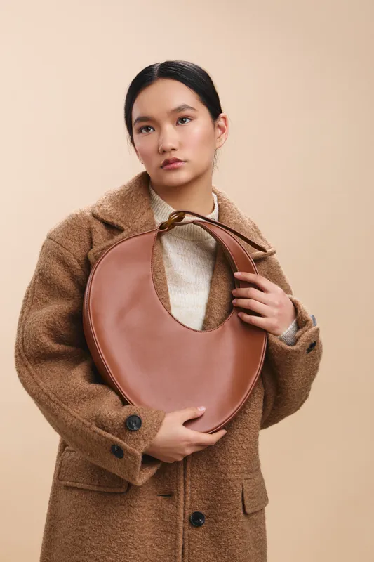 asian woman wearing a tan wool coat holds a round brown handbag against a soft beige backdrop, presenting a chic fashion portrait with calm confidence.