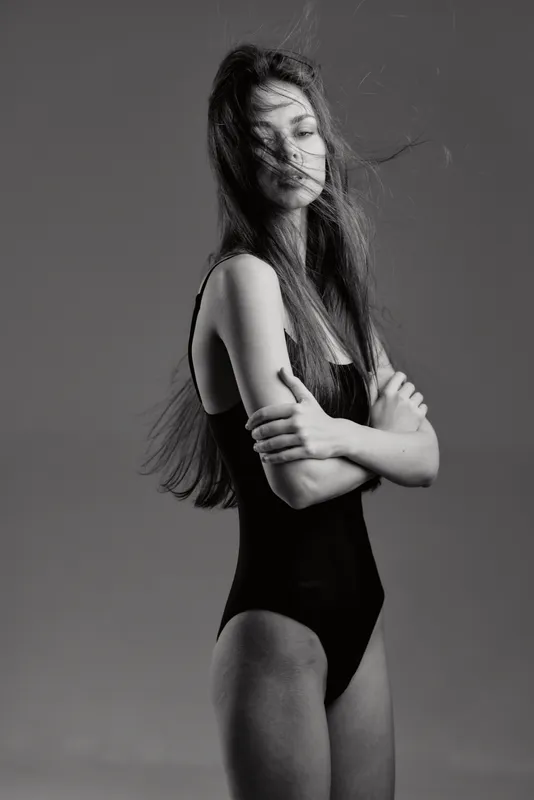 Black and white portrait of a young woman in a black swimsuit with crossed arms and windblown hair, expressing calm and confidence.