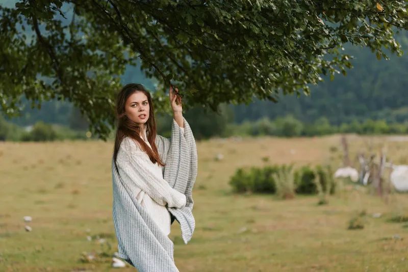 Young woman outdoors wrapped in cozy blanket standing under tree in field, contemplative expression, natural light, casual lifestyle, autumn atmosphere