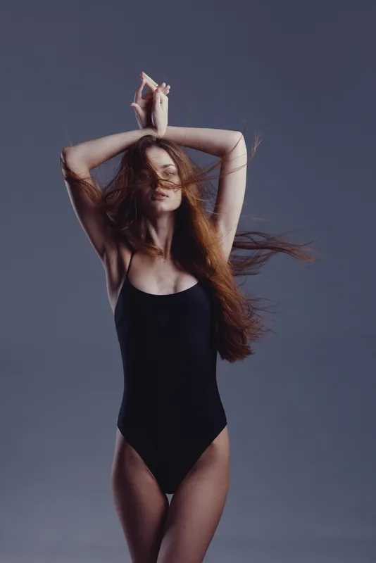 graceful woman posing in black leotard with flowing red hair against gray background