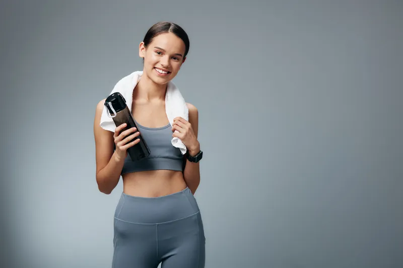 Athletic woman in gym wear with a water bottle and towel draped over her shoulders, posing in a neutral studio. Fitness, sport, health, strength, motivation and active lifestyle.