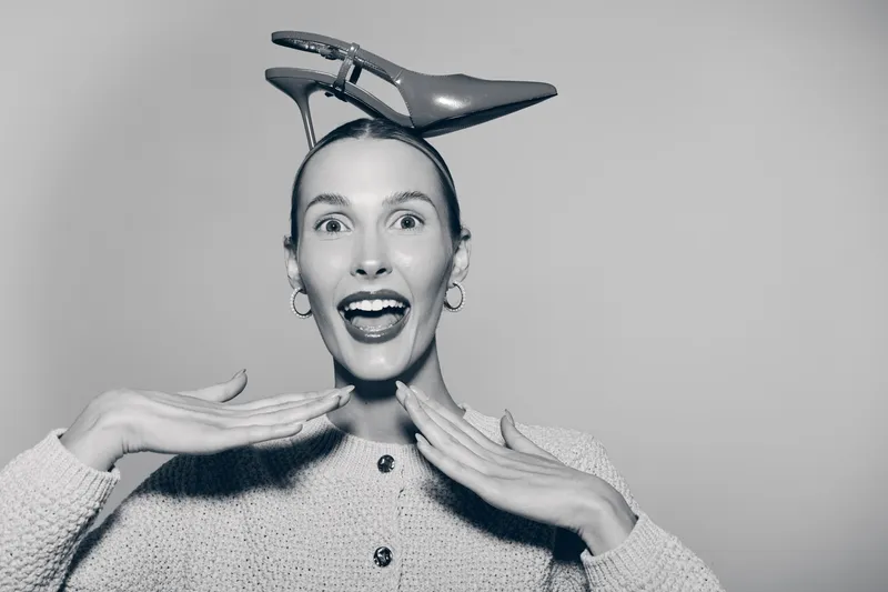 Happy white woman with fair skin and slicked hair holds hands near face with open mouth showing excitement smiling with high heel shoe on head. Studio portrait on plain background.