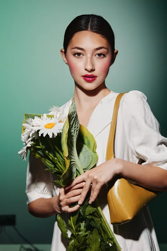 Confident young woman wearing white dress holds a large bouquet of daisies against green background. Soft makeup and red lips create a fresh beauty fashion concept.