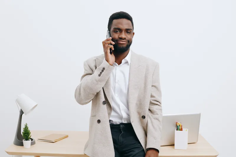 Professional man with beard talking on mobile phone during work in office, dressed in smart casual attire, standing beside desk with laptop, stationery, and plant, white background, business lifestyle