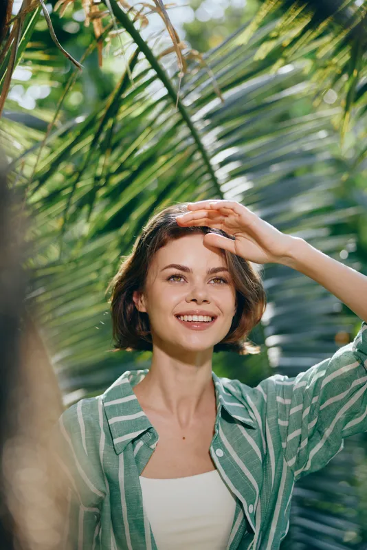 Portrait of a smiling woman shielding her eyes from bright sun among lush tropical greenery, wearing a striped green shirt.