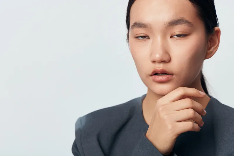 Young Asian woman with hand near chin in casual blazer against plain background, thoughtful expression, closeup portrait showing natural makeup and neat hairstyle for confidence.