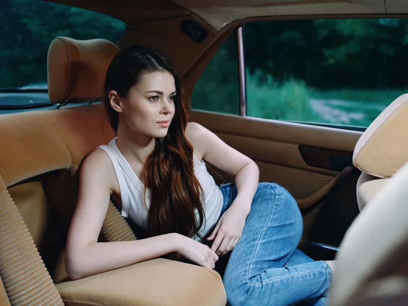 Young woman with long hair in casual outfit relaxing in a vintage car, featuring a natural, serene expression and green outdoor background.