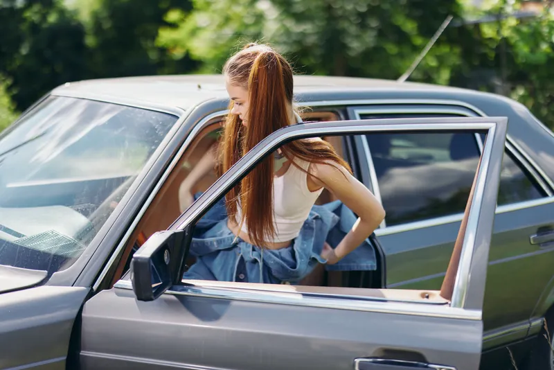 Young woman with long hair stepping out of a vintage car on a sunny day, dressed in casual attire with a denim jacket, evoking a carefree summer vibe.