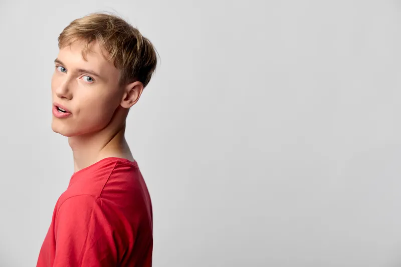 Man in a red shirt glances over the shoulder in a clean studio, minimal background, confident expression, lifestyle fashion shoot with sharp, natural lighting and simple setting