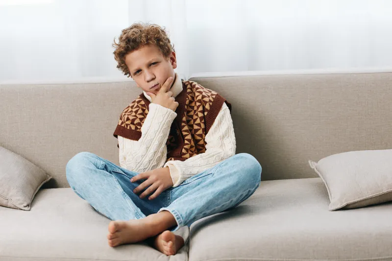 Young boy sits barefoot on a light sofa, wearing a patterned scarf and knitted sweater, gazing thoughtfully with a pensive expression and relaxed posture in a cozy living room.