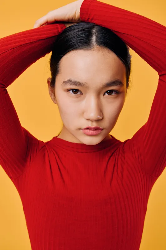 Young woman in red sweater posing with hands behind head against bright yellow background. Portrait of Asian female model with serious expression and casual fashion style indoors.