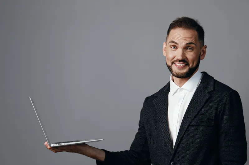 A professional man in business attire holds a laptop, presenting a confident, friendly smile against an isolated colorful background, conveying success and engaging corporate energy.
