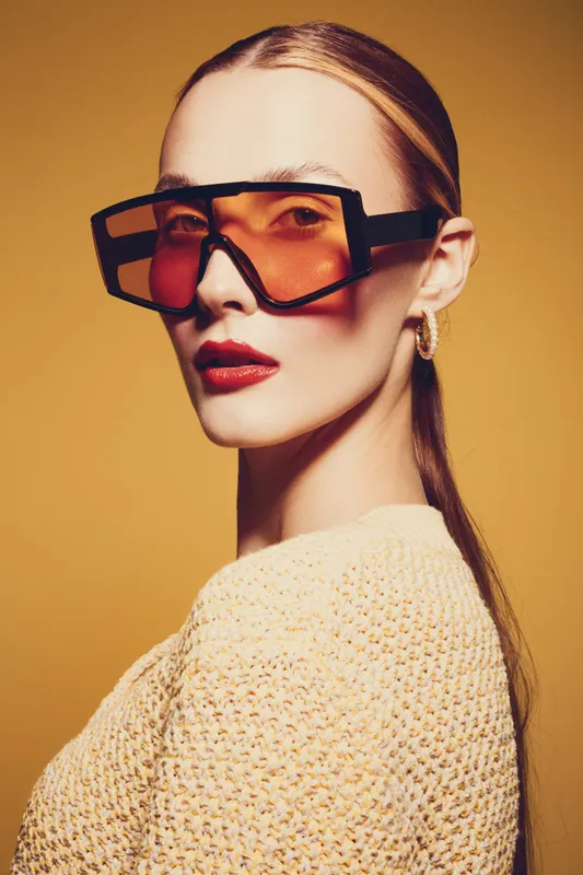 Stylish woman with slicked back hair wears oversized orange sunglasses and knitted sweater against yellow background. Concept of fashion, modern style, accessories, and confident attitude.