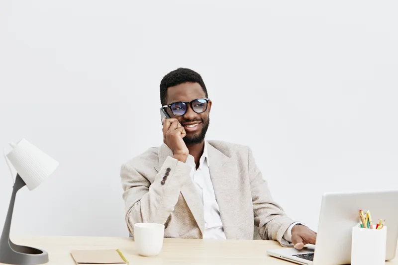 Professional young African American man in beige blazer and white shirt smiling and talking on mobile phone while working at modern office desk with laptop, mug, and stationery isolated on light backg