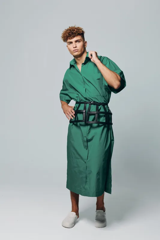 Young man with curly hair wearing green long shirt dress and black belt bag posing in studio with neutral background. Casual fashion, confident model and modern street style outfit.