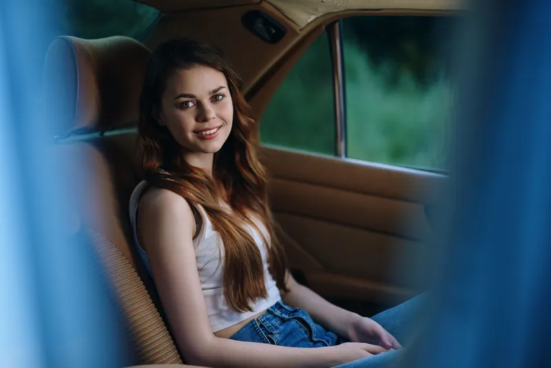 Young Caucasian woman smiling in a car with a relaxed expression, wearing a casual outfit and surrounded by greenery, perfect for lifestyle and travel themes.