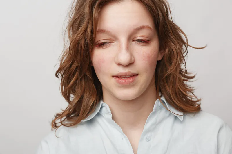Young woman with wavy brown hair and light freckles looks down with a soft expression wearing a light blue shirt. Studio portrait on plain white background with natural lighting.