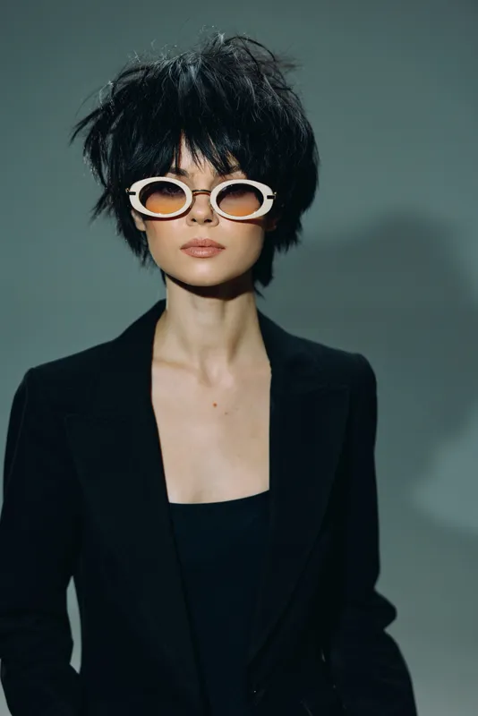 Stylish woman in white sunglasses and black blazer posing with short messy hair on dark background