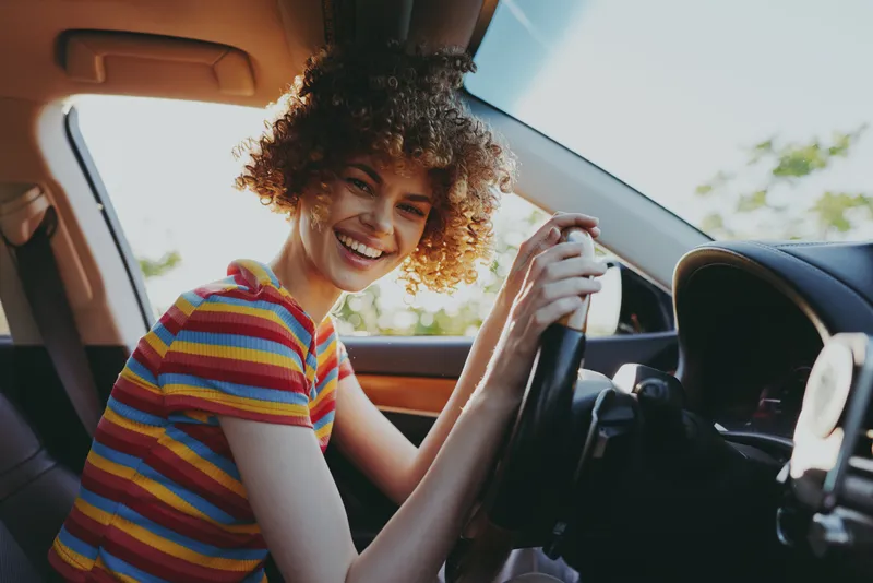 Smiling woman with curly hair driving a car, striped shirt, bright sunlight through the window. A candid lifestyle moment highlighting joy, freedom, and everyday travel in warm color.