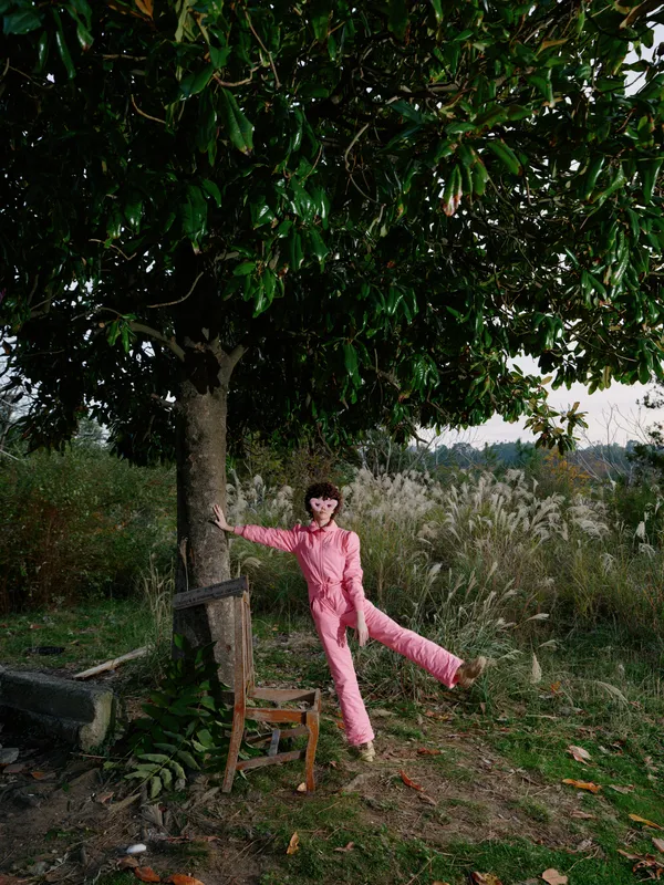 Child in a pink jumpsuit leans against a wooden fence under a leafy tree, posing playfully in a rural setting with tall grasses and distant fields, capturing a bright outdoor lifestyle moment