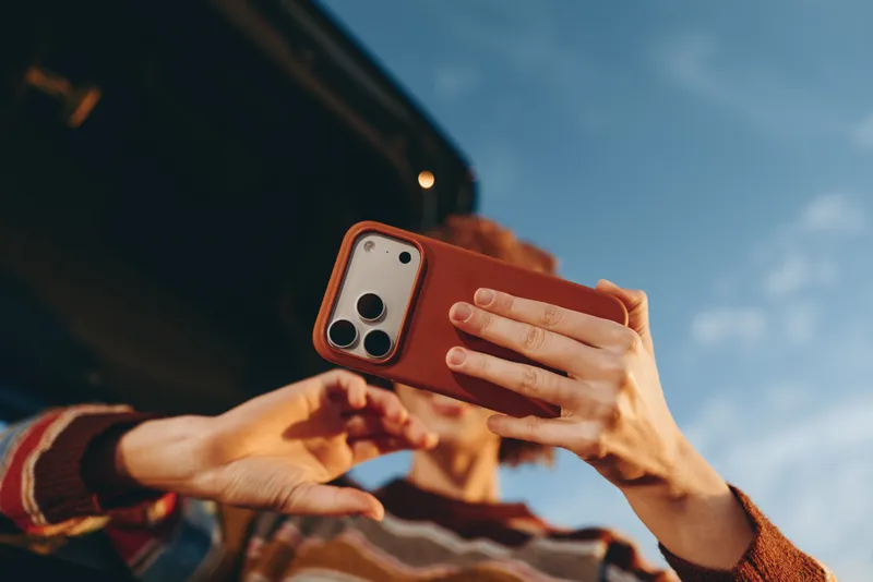 Woman lifestyle outdoors selfie moment, close up hands holding smartphone with orange case, sunny blue sky background, casual streetwear, modern communication, natural light relaxed vibe