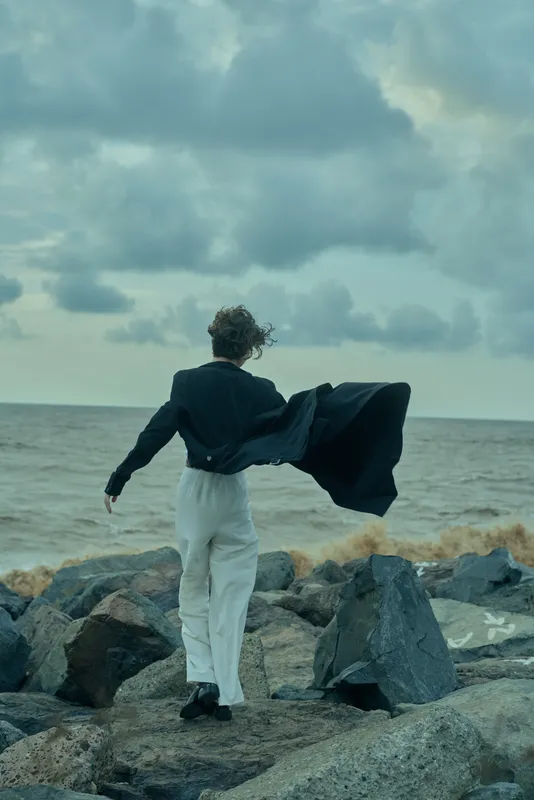 Figure stands on rugged rocks beside the sea, coat billowing in the wind, gazing toward the horizon beneath a dramatic cloudy sky, capturing motion and solitude.