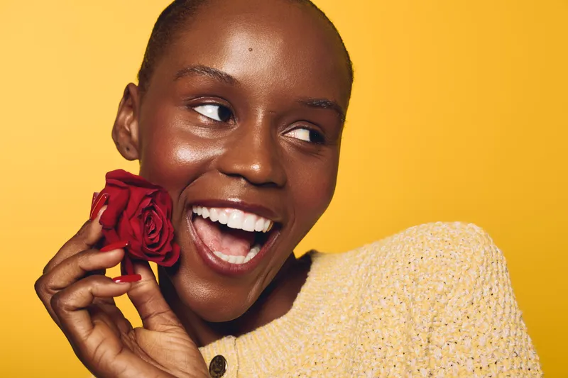 Happy smiling African woman with dark skin holds red rose near face on yellow background. Concept of beauty, joy, natural skin, and positive emotions in studio portrait.