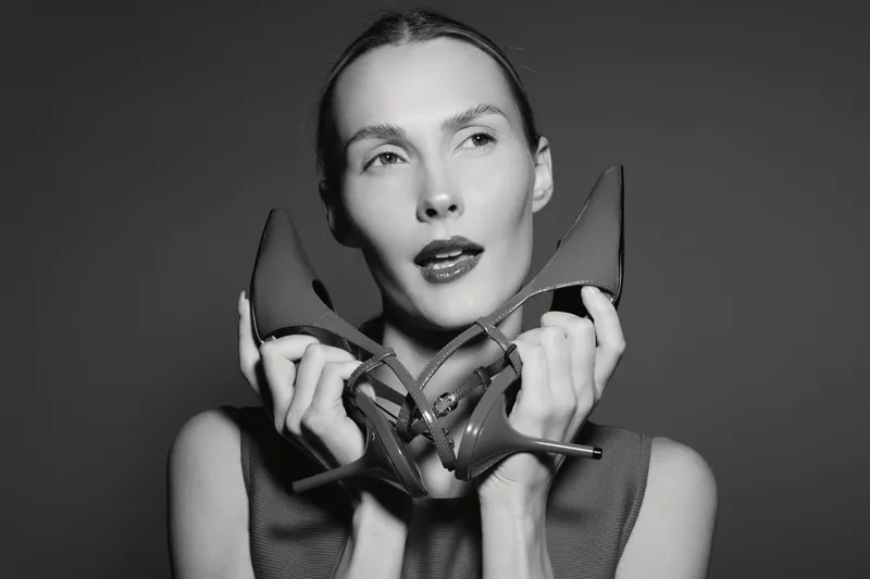 Young white woman with pale skin holds high heel shoes near face looking away in black and white studio portrait. Concept of fashion, style, and beauty with elegant female model.