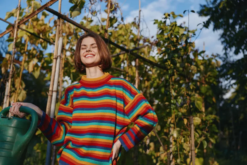 Woman smiling in a rainbow sweater holding a watering can in a sunny garden with green plants, outdoors hobby and casual gardener portrait with cheerful summer mood