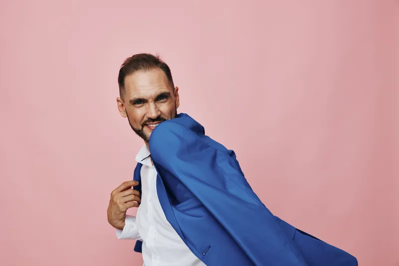 A professional businessman stands against a solid pink background, adjusting his jacket with a confident grin. The pose conveys readiness, mastery, and a strong focus on success.