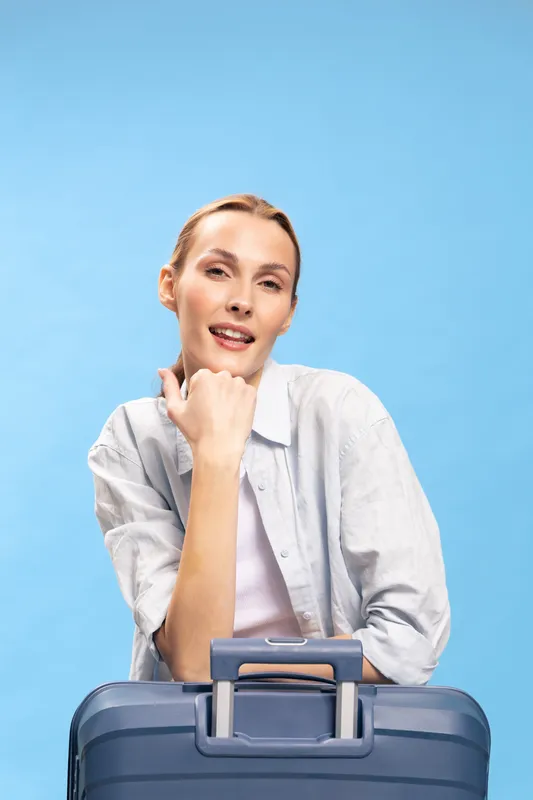 Young woman with light hair and casual shirt poses with blue suitcase on blue background. Travel concept, vacation planning, leisure and tourism. Studio portrait with soft lighting.