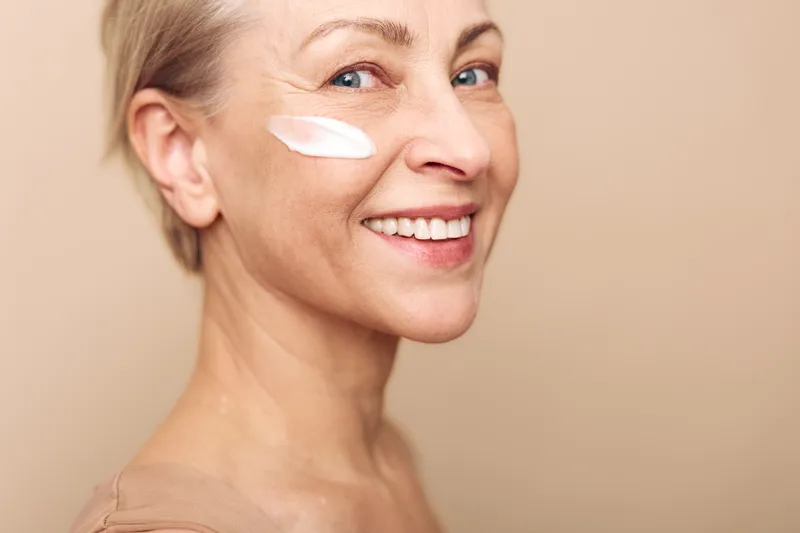 Smiling mature woman with light skin applying moisturizing cream on cheek. Close-up portrait on beige background. Concept of skincare, anti-aging, self-care, natural beauty, and wellness routine.