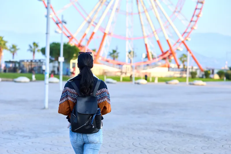 Backpack wearing woman stands in a sunny outdoor plaza, facing a towering ferris wheel at an expansive amusement park, capturing travel and sightseeing vibes, leisure and exploration under clear skies