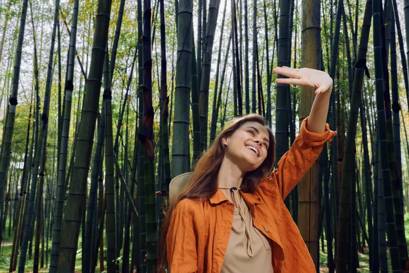 Woman bamboo forest smiling portrait travel scene, joyful young traveler in orange jacket and hat, enjoying sunlight among tall stalks, casual outdoor lifestyle and peaceful nature escape.