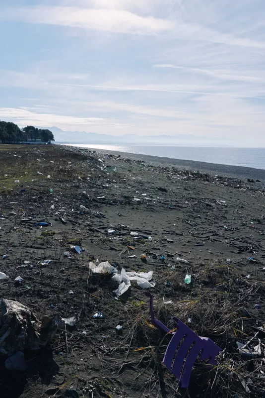 Beach shoreline litter and plastic debris scattered along a rocky coast, trash and waste polluting the calm ocean, wind blown wrappers and bottles dotting the sand and foreshore under cloudy skies
