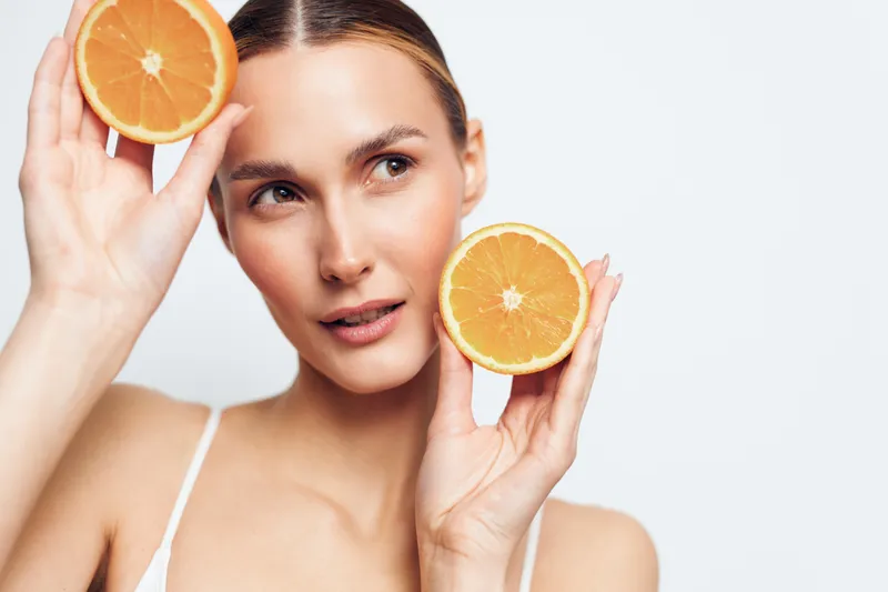 Young woman holding two orange halves near her face, fresh citrus fruit, clear skin, natural beauty, bright lighting, white background, healthy lifestyle concept
