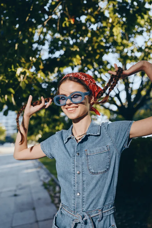 Lifestyle woman in modern boho-western denim street style with film color effect, smiling and playful outdoors in sunny natural environment with green trees background.