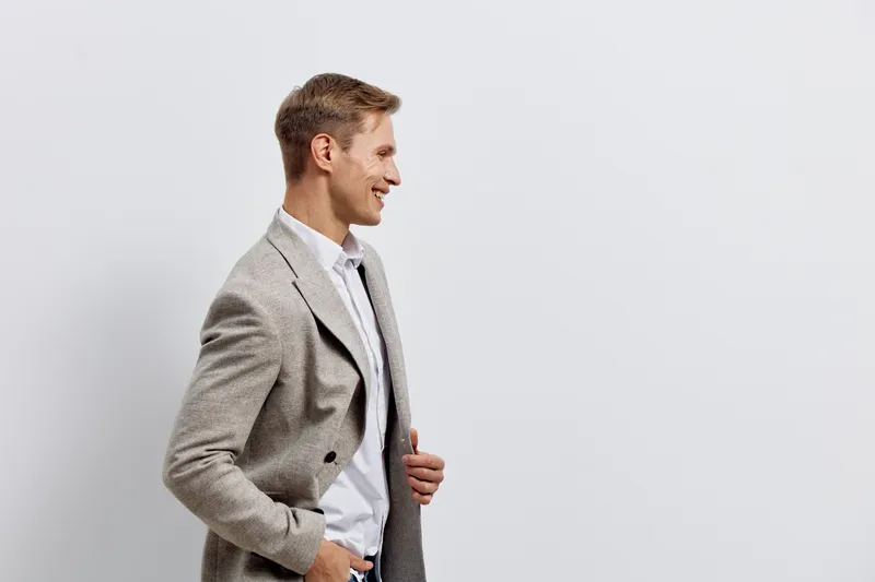 Professional portrait of a smiling young man wearing a gray checkered blazer and white shirt, standing in profile against a plain white background. Studio shot showcasing confident and friendly expres