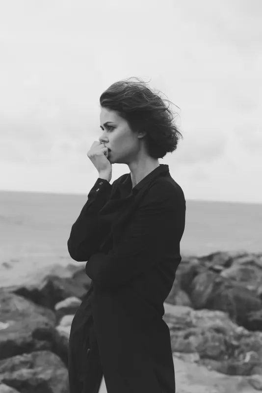 A contemplative woman stands on a rocky coastline, wind in her hair, wearing a dark coat, captured in moody black and white for a poetic, cinematic fashion and reflective mood.