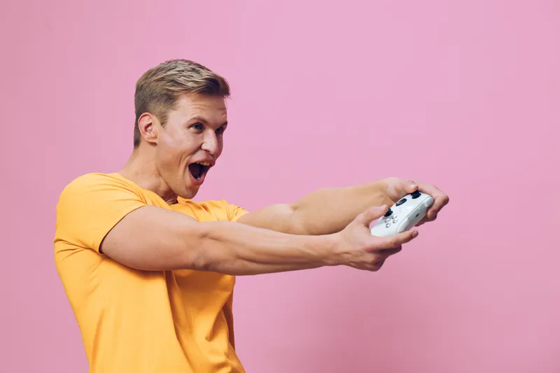 excited young caucasian man in yellow t-shirt holding gamepad playing video game digital detox fun isolated pink background
