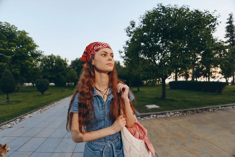 Lifestyle woman in modern boho-western denim street style with a red bandana, casual accessories, and film color effect, posing outdoors in a green park setting.